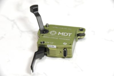 A green, electronic trigger with a flat trigger shoe from MDT. It has a USB-C charging port on the bottom and a right handed safety. White granite is in the background.