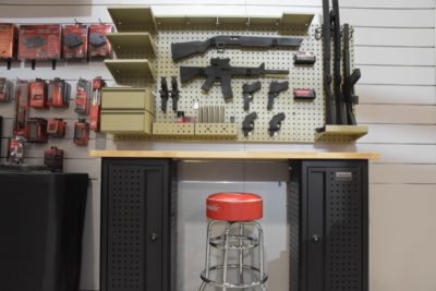 A wall-mounted organization system that uses accessories to secure equipment, firearms, and components to the wall, located above a wooden table top and black cabinets.