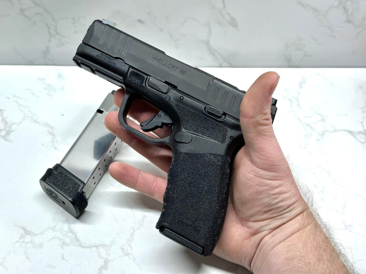 A black handgun lays in the palm of the author's hand. The new Hellcat Pro extended magazine lays in the background against a white marble table top.