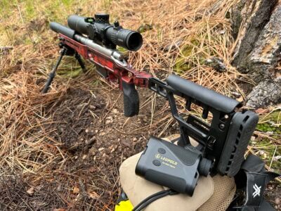 A rangefinder sits on a brown pack next to a red gun in the woods.