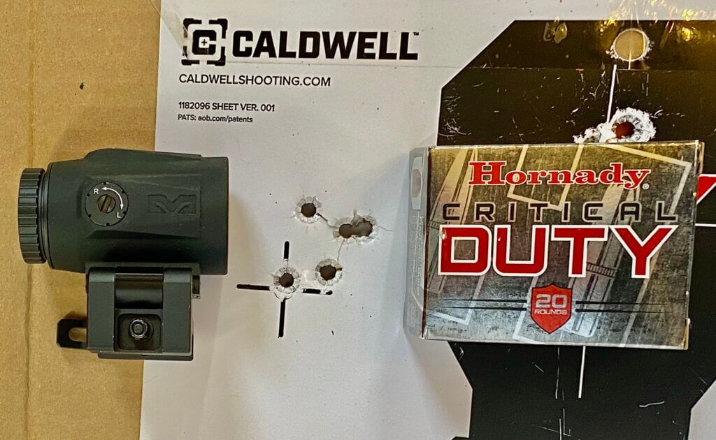 Target with 5 bullet holes next to Hornady Critical duty ammo box and red dot magnifier
