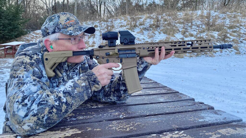Man in camo jacket and hat shooting rifle mounted with Meproguard MMX3 Magnifier outside in the snow