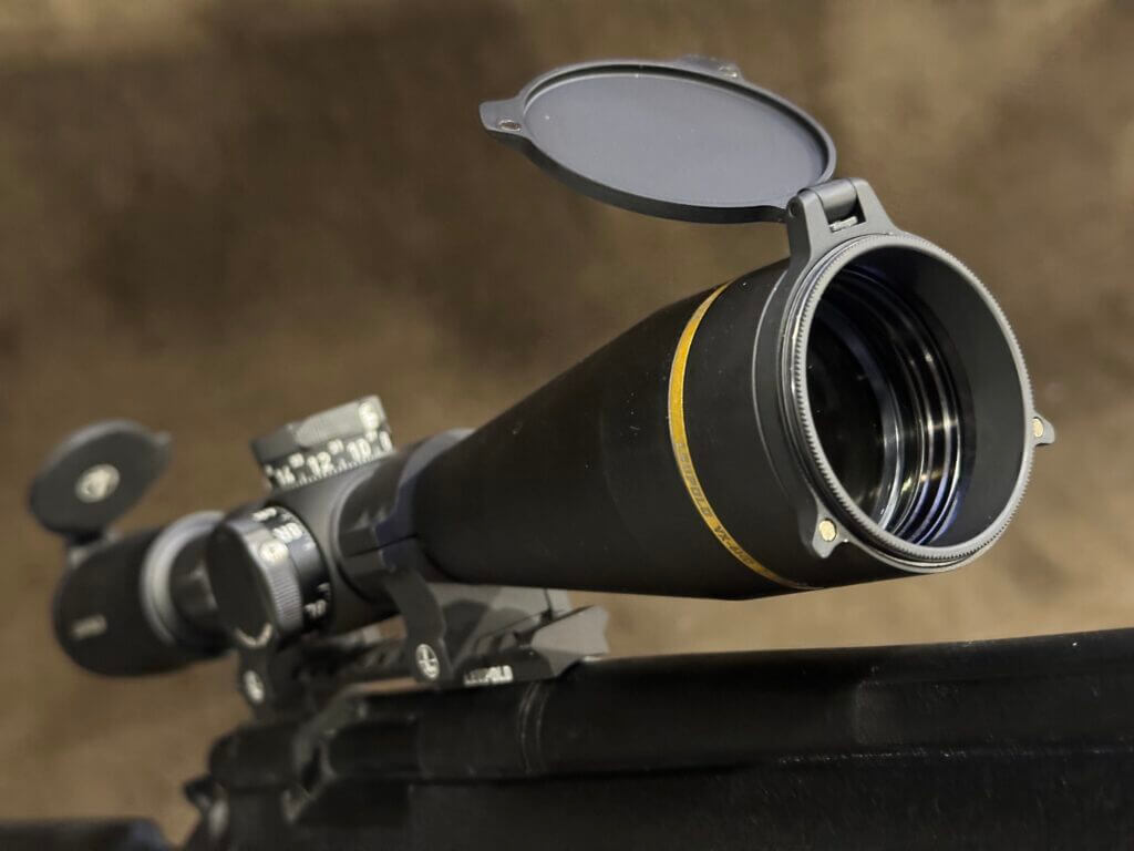 Peering at the rifle scope from the objective lense side, a scope cap is open and the shiny glass lens can be seen.