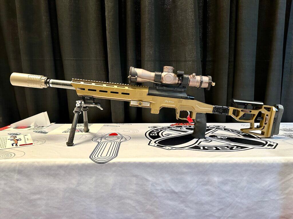 The fully assembled rifle sits on a bipod on a white table, facing to the left.